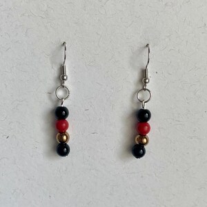 May include: A pair of dangle earrings with a silver hook and a series of beads. The beads are black, red, and gold in color. The earrings are displayed against a white background.