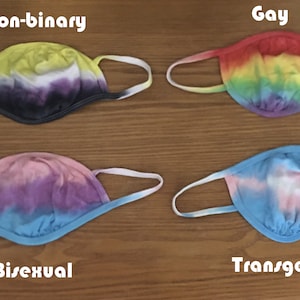 May include: Four face masks with tie-dye patterns representing different sexual orientations. The masks are yellow, red, pink, and blue with white and purple accents. The text "Non-binary", "Gay", "Bisexual", and "Transgender" is written below each mask.