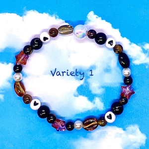 May include: A brown, black, and white beaded bracelet with a variety of charms, including hearts, stars, and a clear iridescent bead. The bracelet is on a blue and white cloudy background. The text "Variety 1" is written on the background.