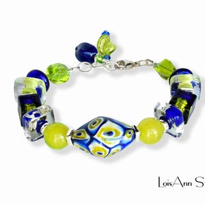 May include: A handcrafted bracelet featuring a variety of colorful glass beads. The bracelet includes blue, green, and yellow beads in various shapes, including a central diamond-shaped bead. The bracelet is connected with a silver clasp.