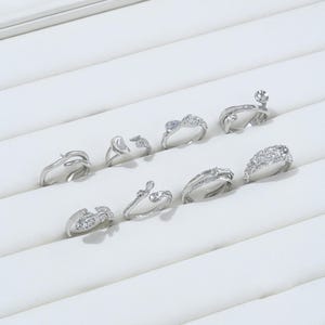 May include: A collection of silver rings displayed on a white surface. The rings feature various designs, including those adorned with clear gemstones and unique shapes. The rings are arranged in a neat, organized manner, showcasing their intricate details.