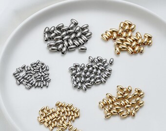 May include: Close-up of various metallic oval beads in silver and gold tones, arranged on a white plate. The beads are small and uniform in shape, ideal for jewelry making or crafting projects. The image shows a variety of bead sizes and quantities.