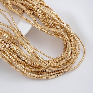 May include: Close-up of multiple strands of gold-colored necklaces. The necklaces feature various bead shapes, including small squares, rectangles, and round beads. The jewelry is arranged in a layered, cascading style against a white background.