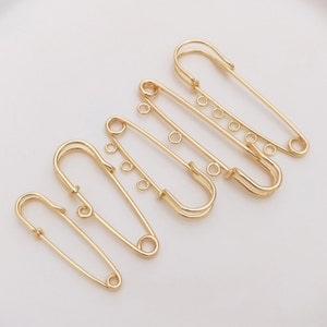 Könnte beinhalten: Ein Satz von fünf goldenen Sicherheitsnadeln in verschiedenen Größen. Die Nadeln sind in einer Reihe angeordnet, wobei die größte Nadel oben und die kleinste Nadel unten ist.