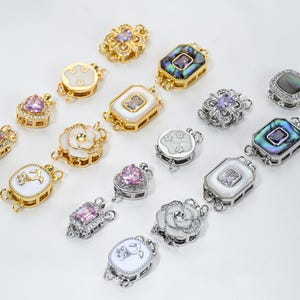 May include: An assortment of decorative jewelry clasps in gold and silver tones. The clasps feature various designs, including floral, heart, and geometric shapes, with inlaid gemstones and mother-of-pearl accents. Some clasps have a white enamel finish.