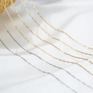 May include: A close-up of several silver and gold chain necklaces with a box chain design. The chains are laid out on a white surface.