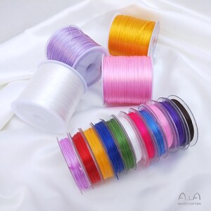 Könnte beinhalten: Eine Auswahl an farbigen elastischen Schnurspulen in verschiedenen Farben, darunter Weiß, Lila, Orange, Rosa, Rot, Gelb, Grün, Blau und Schwarz. Die Spulen sind auf einer weißen Satin-Oberfläche angeordnet, ideal für Schmuckherstellung und Bastelarbeiten.