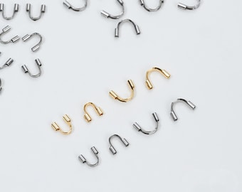 Puede incluir: Una colección de hallazgos de joyería en forma de U, de color plateado y dorado, dispersos sobre una superficie blanca. Los hallazgos tienen un diseño curvo con dos pequeños postes cilíndricos en cada extremo. El texto "Stainless Steel" es visible en la esquina inferior derecha.