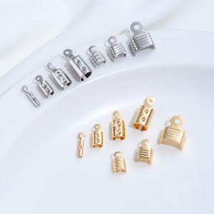 May include: A collection of metal cord end caps in silver and gold tones, arranged in two separate rows by size. Each piece features a cylindrical or wide U-shaped body with a flat top loop for attachment, designed for securing jewellery cords.