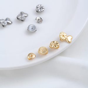 May include: A collection of metallic earring backs in silver and gold tones. The silver backs include circular and heart-shaped designs, while the gold backs feature circular and heart-shaped styles with a textured, scalloped edge design.