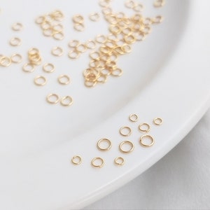 May include: A white plate with a large quantity of small gold jump rings. The jump rings are scattered across the plate, with a few smaller jump rings in the bottom right corner.