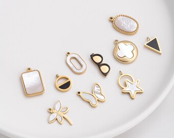 May include: A collection of gold-toned charms with various designs, including a rectangle, oval, clover, star, butterfly, and dragonfly. Some charms feature white mother-of-pearl inlays and black accents. The charms are arranged on a white surface.