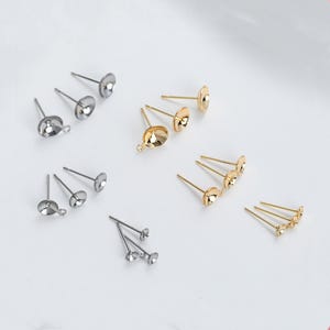 Puede incluir: Un juego de hallazgos de pendientes en oro y plata. Los hallazgos están hechos de metal y tienen un pequeño bucle en la parte superior para sujetar los pendientes. Los hallazgos están dispuestos en filas sobre una superficie blanca.