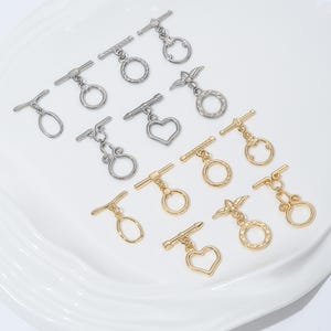 Puede incluir: Un surtido de cierres de palanca plateados y dorados en varios diseños, incluyendo círculos, corazones y formas decorativas. Estos hallazgos de joyería se exhiben sobre una superficie blanca ligeramente texturada.