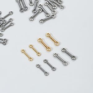 Puede incluir: Un surtido de pequeños conectores de joyería en tonos plateados y dorados. Los conectores tienen un centro de alambre retorcido y bucles circulares en cada extremo. Algunos están dispersos, otros están dispuestos sobre una superficie blanca.