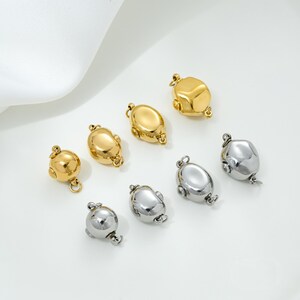 May include: An assortment of gold and silver-toned jewelry clasps. The clasps have a rounded, organic shape with small loops for attaching to jewelry pieces. The clasps are displayed on a white surface.