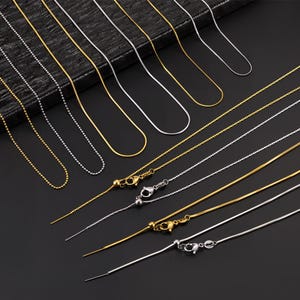 Könnte beinhalten: Eine Auswahl an goldenen und silbernen Halsketten, die auf einem dunklen Hintergrund präsentiert werden. Die Halsketten weisen verschiedene Designs auf, darunter Kugelketten, Schlangenkettchen und feine Panzerketten. Einige haben Verschlüsse, andere sind einfache Anhänger.