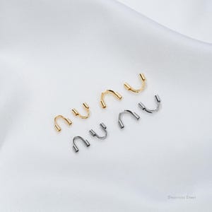 Könnte beinhalten: Eine Auswahl an U-förmigen Schmuckteilen aus Edelstahl in Gold- und Silberfarben. Die Schmuckteile, mit kleinen zylindrischen Enden, sind für die Schmuckherstellung bestimmt. Sie liegen auf einer weißen, strukturierten Oberfläche.