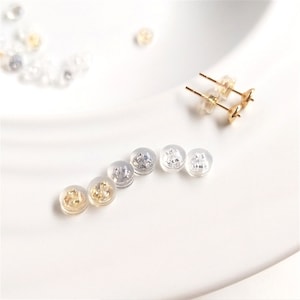 Peut inclure: Diverses attaches de boucles d'oreilles et boucles d'oreilles à clous dorées sur une surface blanche. Les attaches sont transparentes, dorées et argentées. Les boucles d'oreilles ont des tiges dorées et des dos ronds.