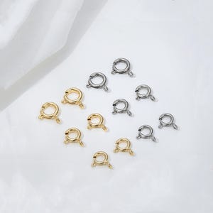 Puede incluir: Un conjunto de 10 anillos de salto dorados y plateados, también conocidos como cierres de langosta, para la elaboración de joyas. Los anillos están dispuestos en dos filas sobre una superficie blanca.