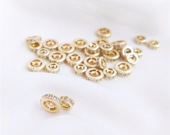 May include: A pile of gold-toned metal spacer beads with clear rhinestones. The beads are round and have a raised edge. There are two beads in the foreground that are separated from the pile.