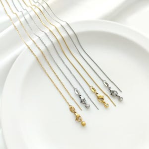 May include: Six different chain styles in gold and silver, each with a lobster clasp closure. The chains are arranged in a row on a white surface.