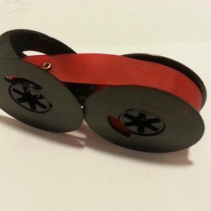 May include: A red typewriter ribbon spool with black plastic spools. The ribbon is wound around the spools.