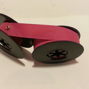 May include: A pink typewriter ribbon on two black spools. The ribbon is wound around the spools and is ready to be used in a typewriter.