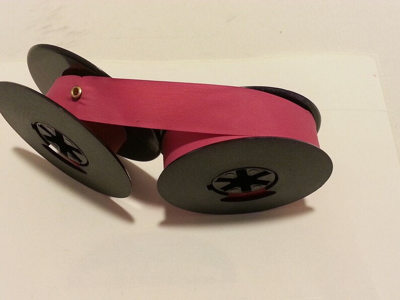 May include: A pink typewriter ribbon on two black spools. The ribbon is wound around the spools and is ready to be used in a typewriter.