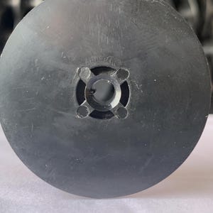 May include: A black, circular plastic object with a central hole and four smaller holes around it. The object has a slightly textured surface and appears to be a component of a larger mechanism or device.