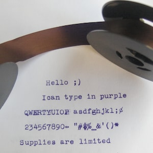 May include: Close-up of a brown typewriter ribbon spool with black plastic spools. The ribbon is threaded through the spools. The text "Hello ;)" is typed in purple ink on a white sheet of paper. The text "I can type in purple" is also typed in purple ink. The text "QWERTYUIOR asdfghjkl;" is typed in purple ink. The text "234567890- "#%&'()*" is typed in purple ink. The text "Supplies are limited" is typed in purple ink.