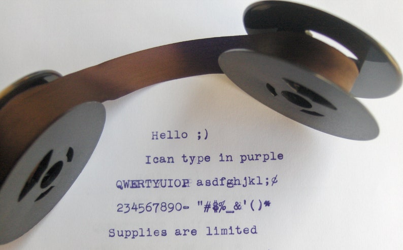 May include: Close-up of a brown typewriter ribbon spool with black plastic spools. The ribbon is threaded through the spools. The text "Hello ;)" is typed in purple ink on a white sheet of paper. The text "I can type in purple" is also typed in purple ink. The text "QWERTYUIOR asdfghjkl;" is typed in purple ink. The text "234567890- "#%&'()*" is typed in purple ink. The text "Supplies are limited" is typed in purple ink.
