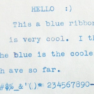 May include: Typewritten text on a white piece of paper that reads "HELLO :) This a blue ribbon and it is very cool. I think that the blue is the coolest color we have so far. "#%&'()*234567890-"