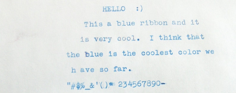 May include: Typewritten text on a white piece of paper that reads "HELLO :) This a blue ribbon and it is very cool. I think that the blue is the coolest color we have so far. "#%&'()*234567890-"