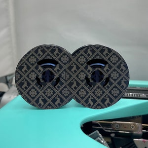 May include: Two black round wooden coasters with a brown and white geometric pattern featuring reindeer and snowflakes. The coasters are on a turquoise surface.