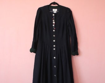 Könnte beinhalten: Ein langärmeliges schwarzes Kleid mit Knopfleiste und einer Reihe von dekorativen Knöpfen. Das Kleid hat einen leicht ausgestellten Rock und einen grünen Saum an den Ärmeln.
