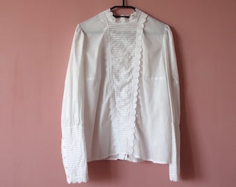 Pode incluir: Uma blusa branca com gola alta e acabamento em renda pendurada em um cabide preto contra uma parede rosa.