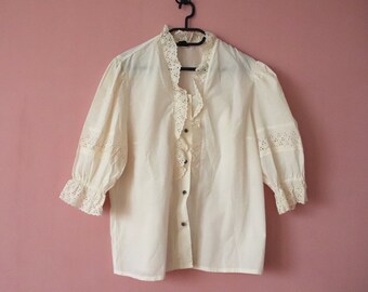Pode incluir: Uma blusa branca com acabamento em renda e fechamento com botões na frente. A blusa está pendurada em um cabide preto contra uma parede rosa.