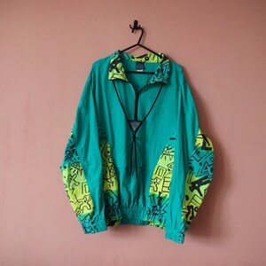 May include: A teal blue windbreaker jacket with black and neon green geometric patterns on the sleeves. The jacket has a zip closure and a drawstring at the waist.