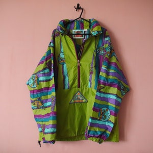 May include: A green, purple and blue patterned jacket with a zip closure and a small logo patch on the front. The jacket has a drawstring hood and elastic cuffs.