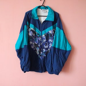 May include: A blue, turquoise and white windbreaker jacket with a floral pattern on the front. The jacket has a zip closure and a stand-up collar.