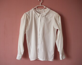 Pode incluir: Uma blusa branca de manga comprida com colarinho de renda e fecho de botões na frente.
