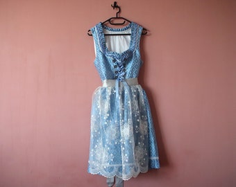 May include: A blue and white dirndl dress with a white lace overlay and a blue ribbon around the waist. The dress is hanging on a black hanger against a pink wall.