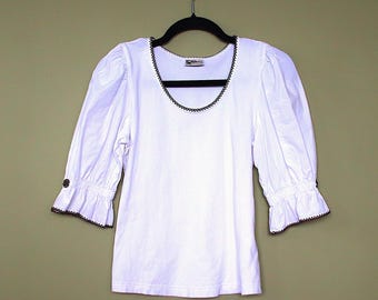 Pode incluir: Camisola branca de manga curta com acabamento preto em volta do decote e punhos. A camisa tem uma manga ligeiramente bufante.