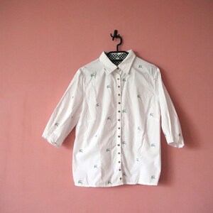 May include: A white button-down shirt with a small, embroidered design. The shirt has a collar and long sleeves.