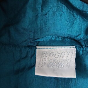 May include: A white tag with the text "SPORT GERMANY" and a logo of two stylized feet. The tag is sewn onto a teal blue jacket.