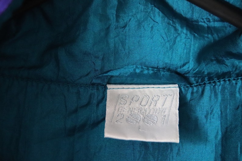 May include: A white tag with the text "SPORT GERMANY" and a logo of two stylized feet. The tag is sewn onto a teal blue jacket.