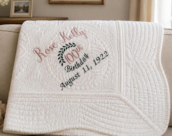 May include: A white quilted blanket with embroidered text reading "Rose Kelly 100th Birthday August 11, 1922". The blanket is draped over a beige sofa. The embroidery is in pink and black.