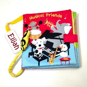 May include: A red, yellow, blue, and green fabric book with the title "Musical Friends" and a name tag that says "Elijah". The book features a monkey playing drums, a dog playing piano, an elephant playing trumpet, and a duck playing saxophone.