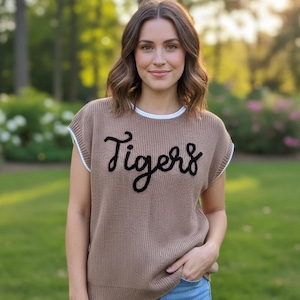 May include: A light brown sleeveless knit sweater vest featuring a crew neckline with white trim and the word Tigers embroidered in black cursive script across the chest, paired with blue denim jeans.